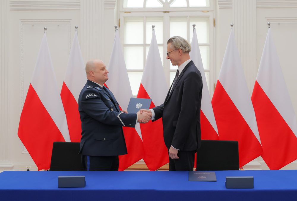 dwóch mężczyzn w garniturach podaje sobie rękę po podpisaniu porozumienia o współpracy, w tle flagi państwowe