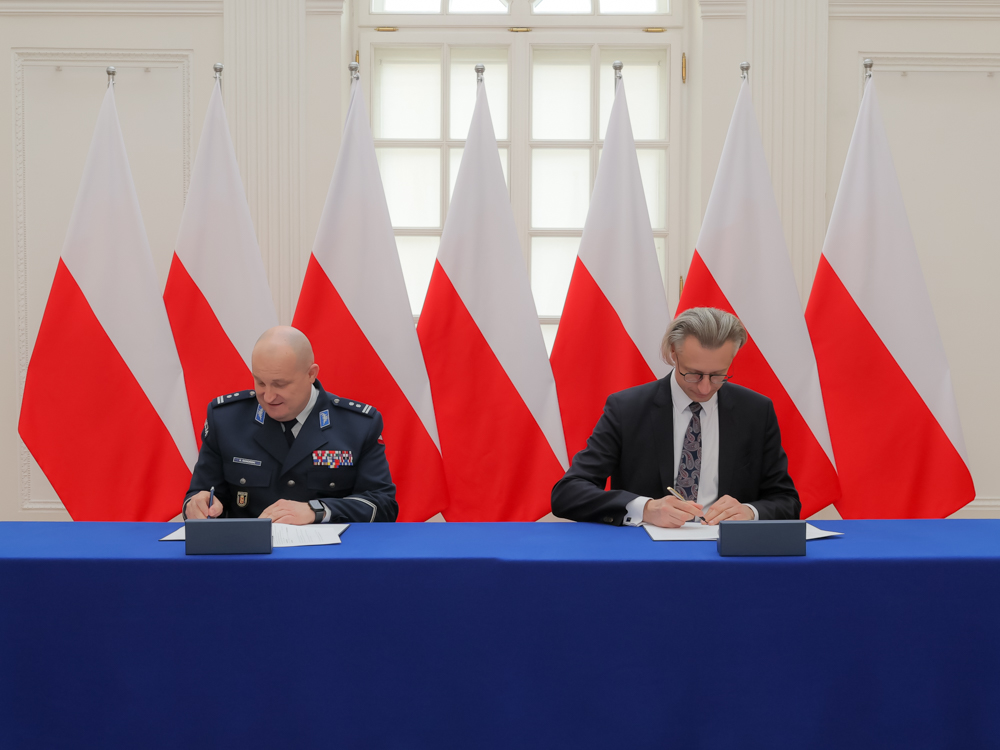 dwóch mężczyzn w formalnych ubraniach podpisuje dokumenty przy stole prezydialnym