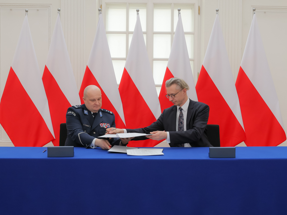 dwóch mężczyzn w formalnym ubraniu przy stole prezydialnym podpisuje dokumenty